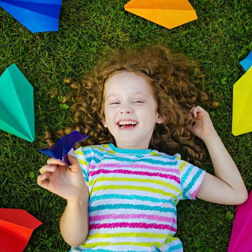 Happy,Laughing,Girl,Throwing,Paper,Airplane,In,Green,Grass,At